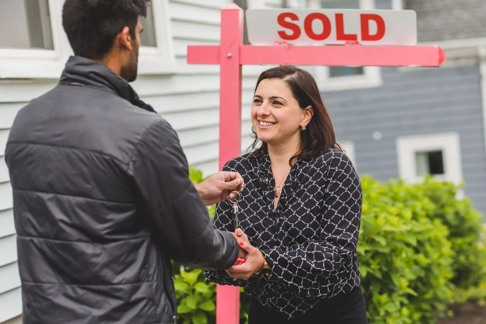 Buyer and seller shaking hands as the house keys are handed over in front of a SOLD sign